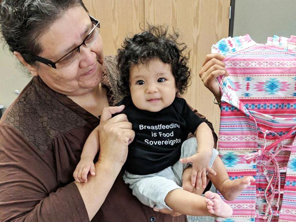 Baby in a shirt that says Breastfeeding is Food Sovereignty being held by a woman