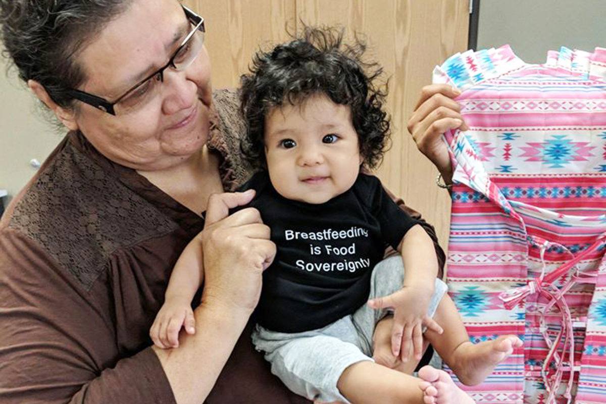 Baby in a shirt that says Breastfeeding is Food Sovereignty being held by a woman